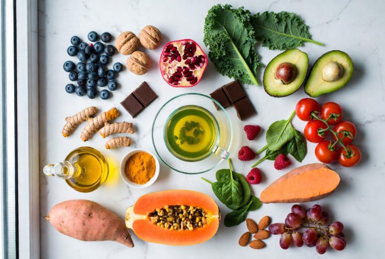 Flat lay of 20 best anti-aging foods including blueberries, walnuts, pomegranate, kale, avocado, green tea, turmeric and more on white marble