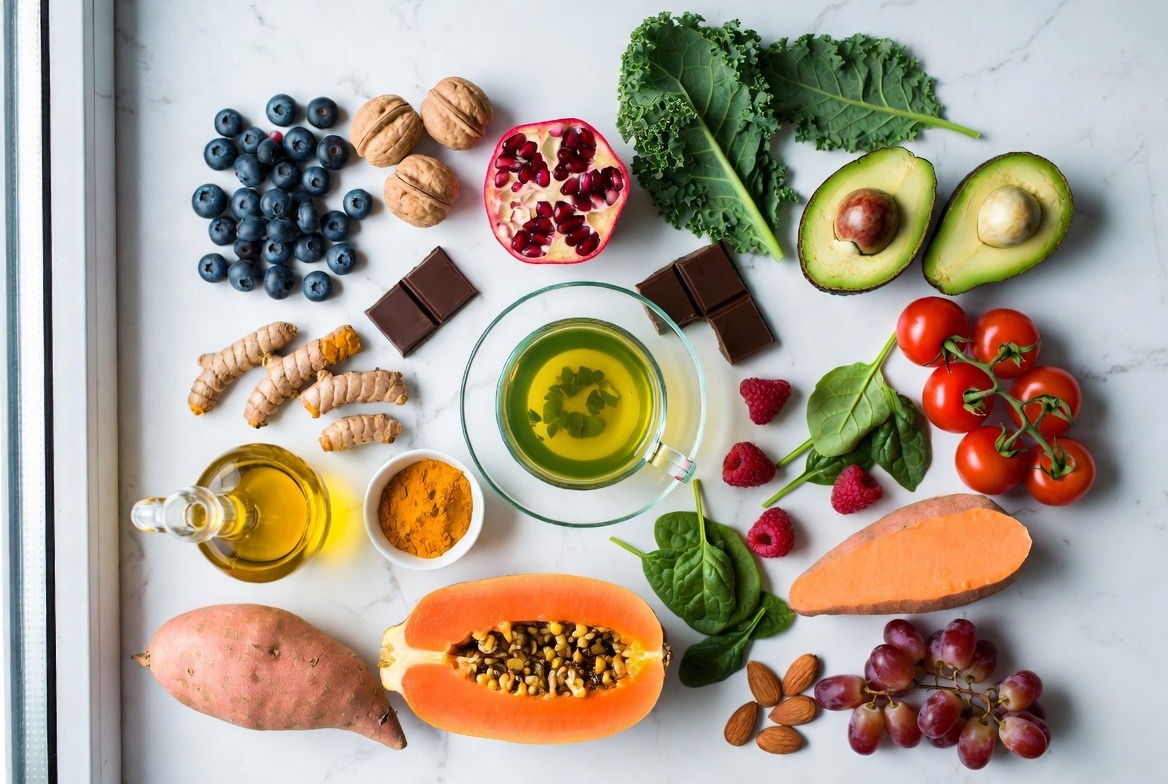 Flat lay of 20 best anti-aging foods including blueberries, walnuts, pomegranate, kale, avocado, green tea, turmeric and more on white marble